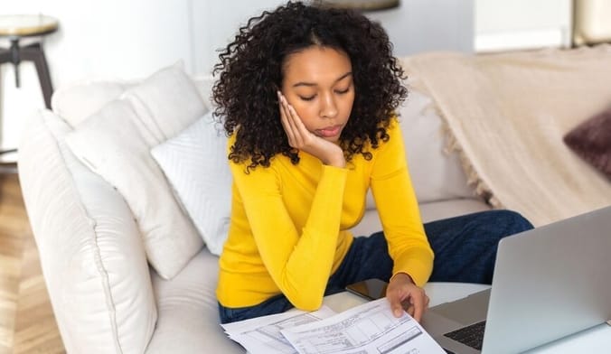A woman with curly dark hair wearing a yellow sweater sits on a white couch in front of an open laptop looking at bills