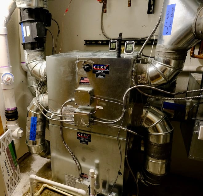 A silver-colored boxy machine, about four feet tall, connected to multiple ducts in a utility closet.