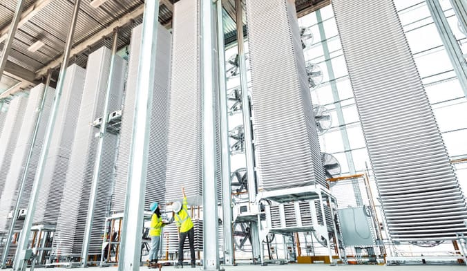 Two workers in safety gear stand next to very large stacks of metal trays in an industrial facility