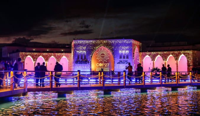People stand on a bridge over a small pond looking at a building with the words Cop 27