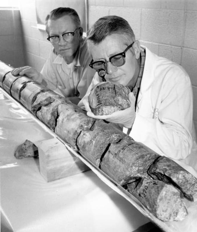 A black and white photo of two men wearing eyeglasses and lab coats looking closely at a piece of granite