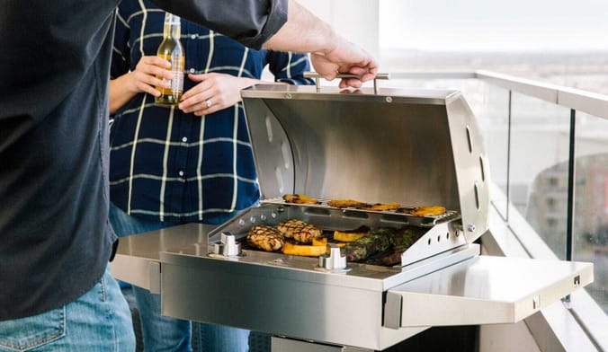 Two people on a balcony in front of a large silver metal grill that has food with sear marks on the 2 levels of grates.
