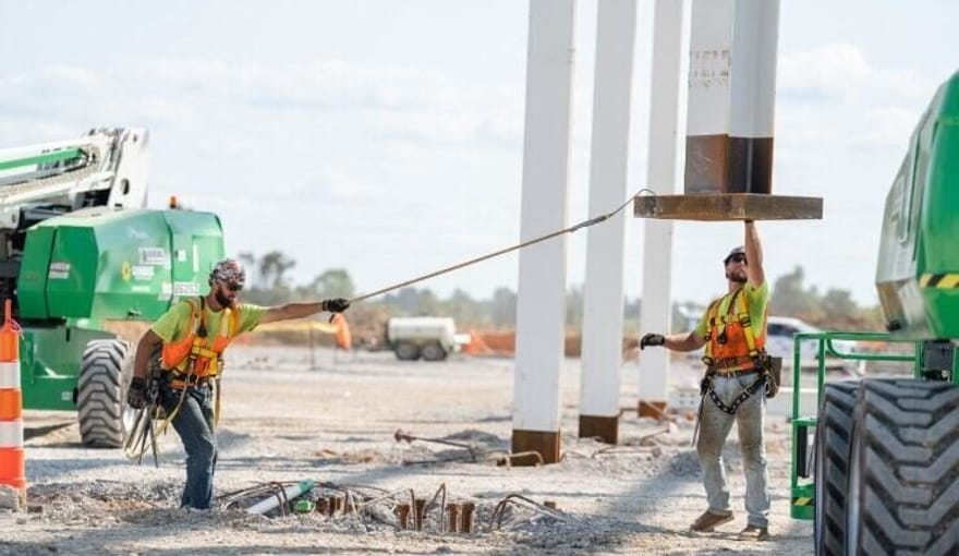 Two men in safety gear and hard hats perform construction work surrounded by heavy equipment
