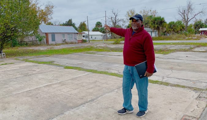 A Black man in a red pullover and jeans stands on a large cement pad and points toward a modest home in the distance.
