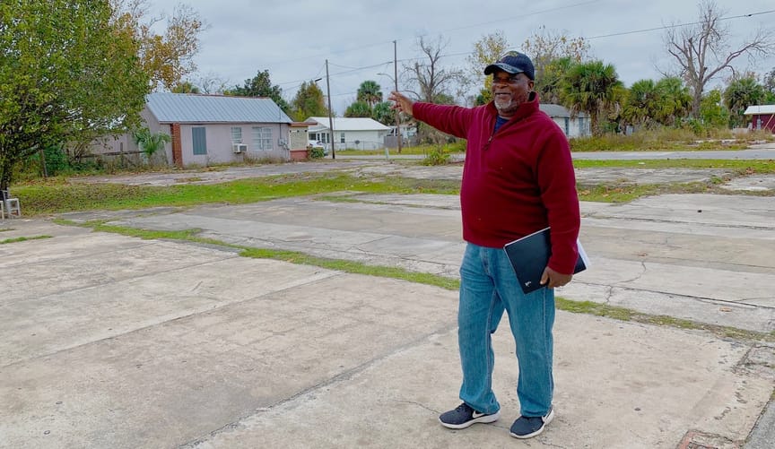 A Black man in a red pullover and jeans stands on a large cement pad and points toward a modest home in the distance.
