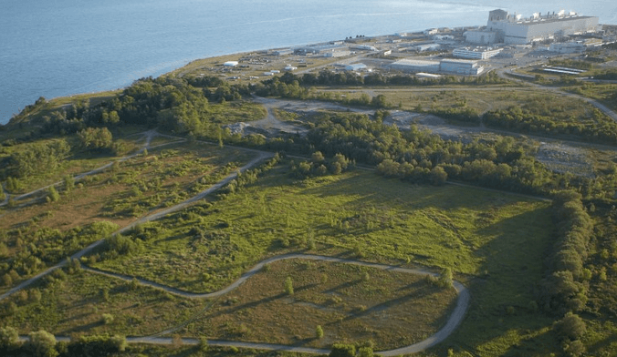 A large industrial facility sits on a verdant hilltop next to a body of water