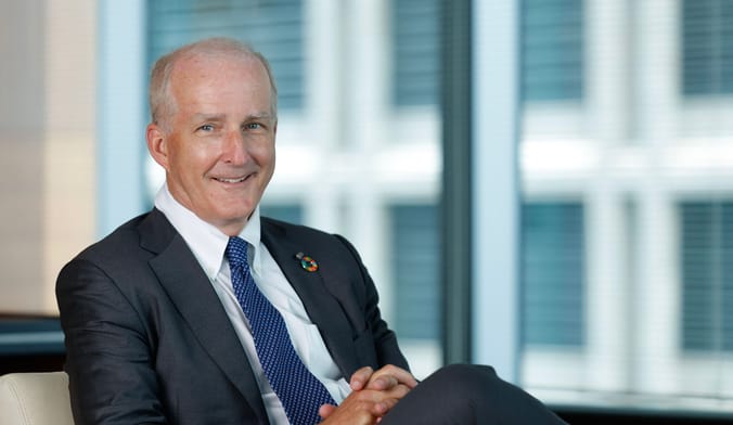 A white man with white hair wearing a dark suit smiles while sitting at a desk in an office