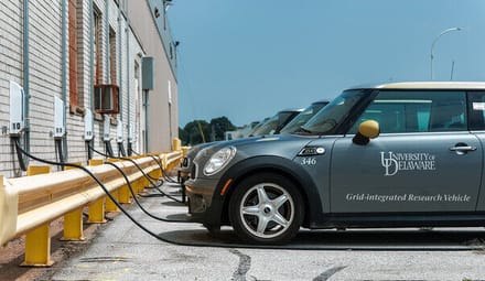 A row of electric vehicles with University of Delaware labels on their doors are connected to chargers attached to a building