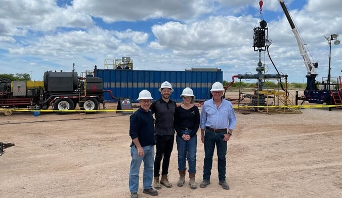 Four people wearing white hard hats stand on a dusty field with drilling equipment in the background.