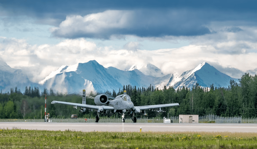 A military plane sits on a runway surrounded by snowy mountains and pine trees