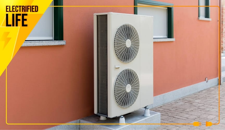 A heat pump's outdoor component, which looks like a tall box with fans, stands outside a building a salmon pink exterior.