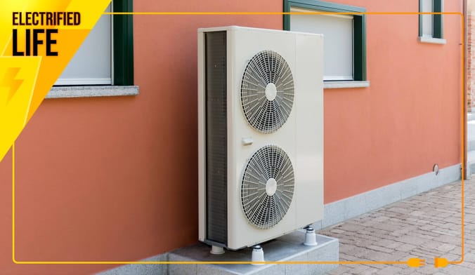A heat pump's outdoor component, which looks like a tall box with fans, stands outside a building a salmon pink exterior.