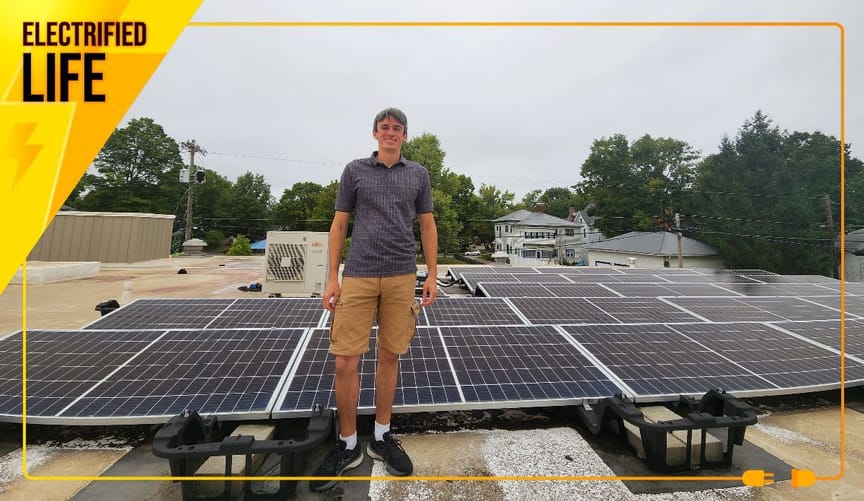 Man in shorts and polo shirt standing in from of 5 rows of solar panels on top of a building roof.