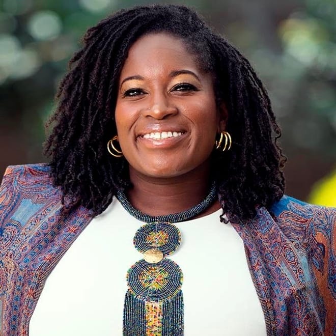 A smiling Black woman with shoulder-length dark curly hair wearing metallic hoop earrings and a multicolored top