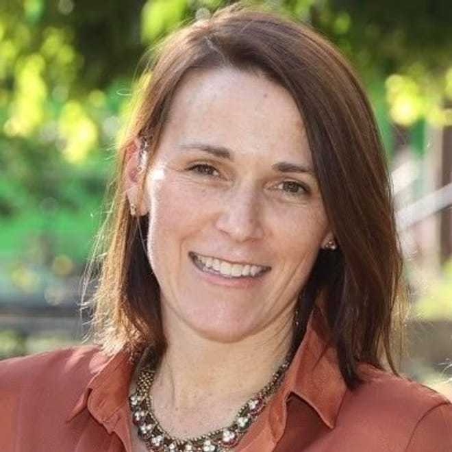 A smiling woman with light skin tone and above shoulder length reddish brown hair wearing a copper top and metallic necklace