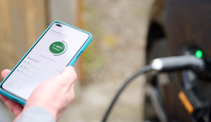 ev.energy app displayed on smartphone held by driver, with EV charging plug in the background
