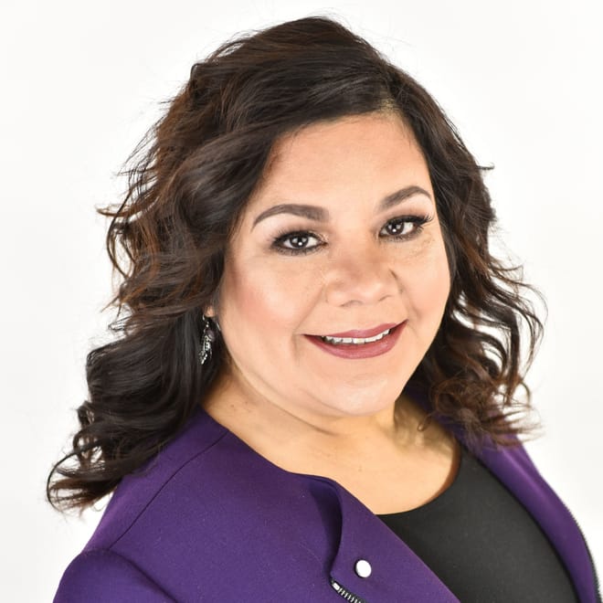 A smiling woman with medium skin tone and shoulder-length dark, wavy hair wearing a purple blazer and black top