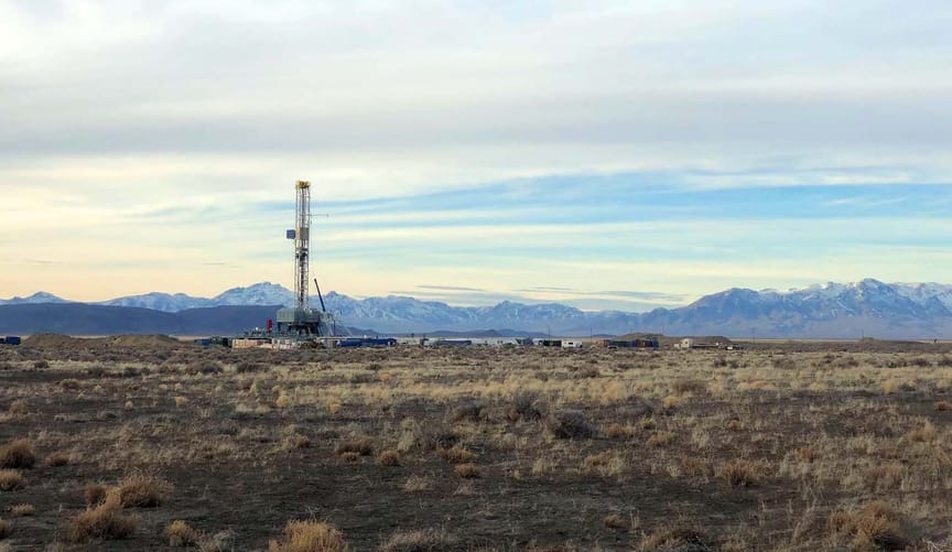 A drilling rig stands tall on a flat landscape with blue mountains in the background