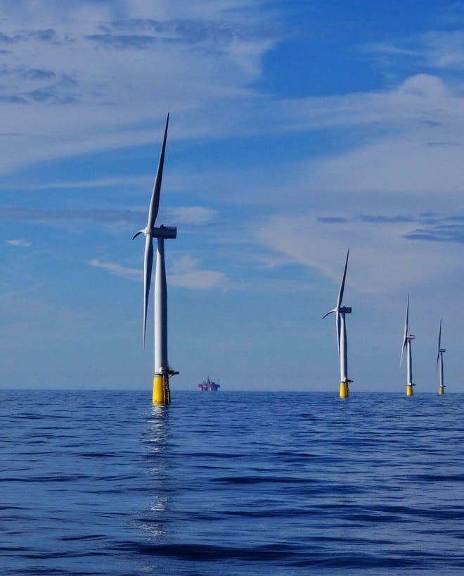 four offshore floating wind turbines in the ocean on a brilliantly sunny day. A ship is seen far in the distance.