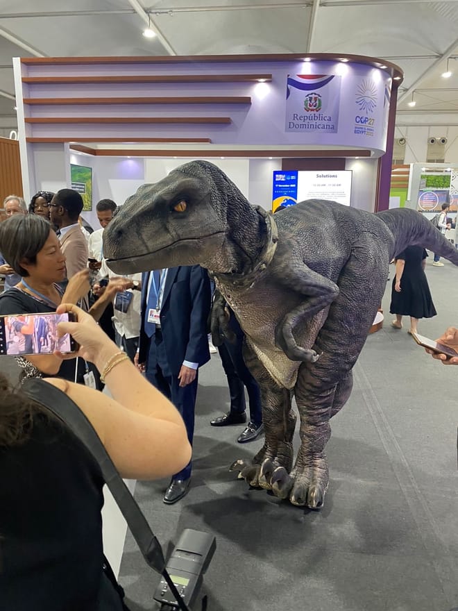people stand around a life-sized model of a dinosaur. Some are taking photos of it.