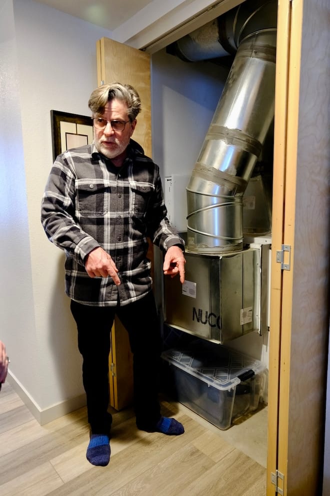 Man with a swoop of silvery hair points downward next to large boxy, metal appliance with vent coming out of it.