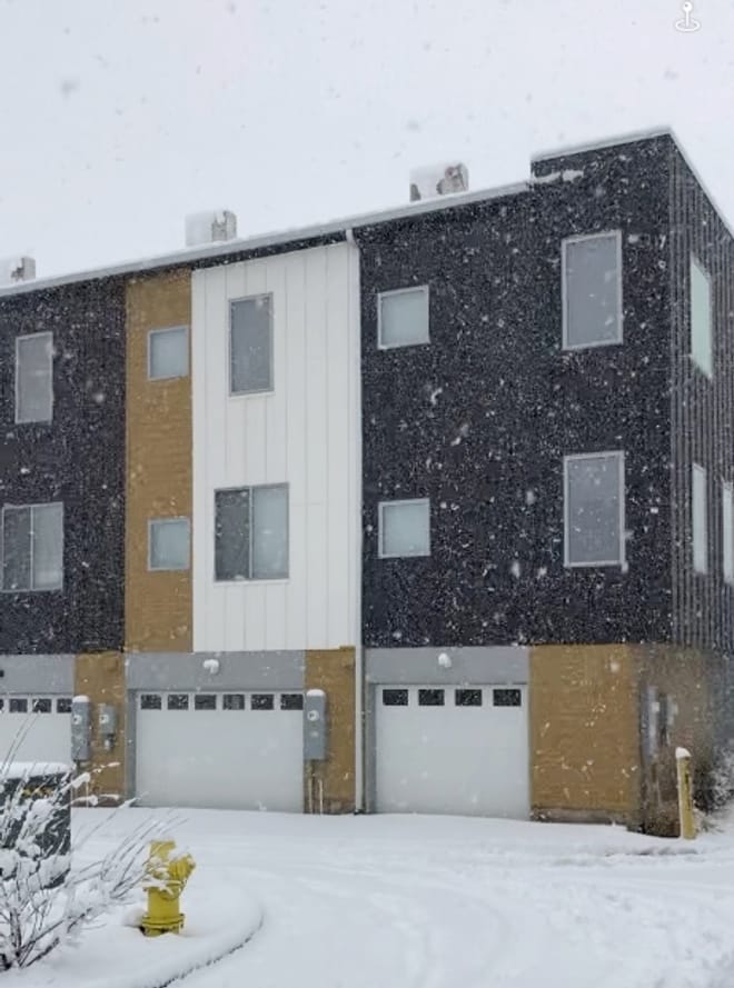 Three-story attached townhomes painted dark blue, white, and tan on a snowy day with rectangular heat-pump units on the roof.