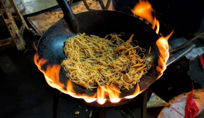 Noodles cook in a wok over a gas-powered flame.
