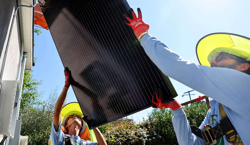 Two people hoist a solar panel onto the roof of a house for a rooftop solar installation.