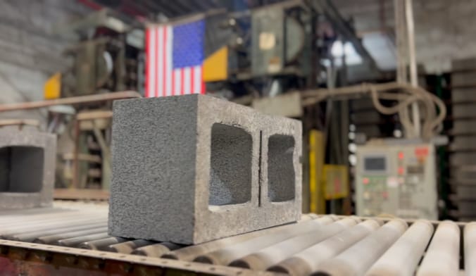 A rectangular gray concrete block on a factory conveyer belt. A U.S. flag draped on equipment is seen in the background.