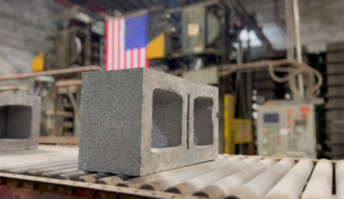 A rectangular gray concrete block on a factory conveyer belt. A U.S. flag draped on equipment is seen in the background.
