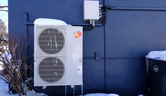 A snow-capped grey Mitsubishi air-source heat pump with circular grills installed outside a blue home in Golden, Colorado.