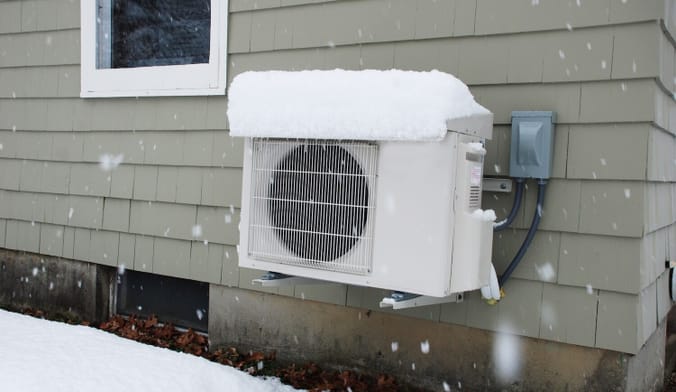 White rectangular appliance equipment - a heat pump - under a cap of snow. The heat pump is mounted to the side of a house.