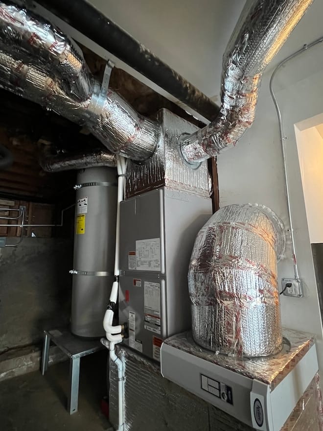 In a utility room, grey water heater tank tank, silver-colored ducts run to large grey metal boxy appliance - blower.