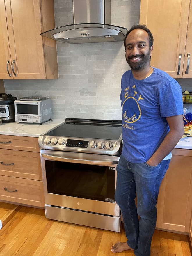 A man with short brown hair and a beard wearing a blue t-shirt and jeans smiles next to a shiny silver range and vent hood