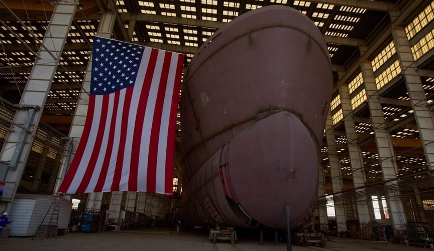 The bow and keel of a large red ship are seen inside a shipyard next to a giant American flag