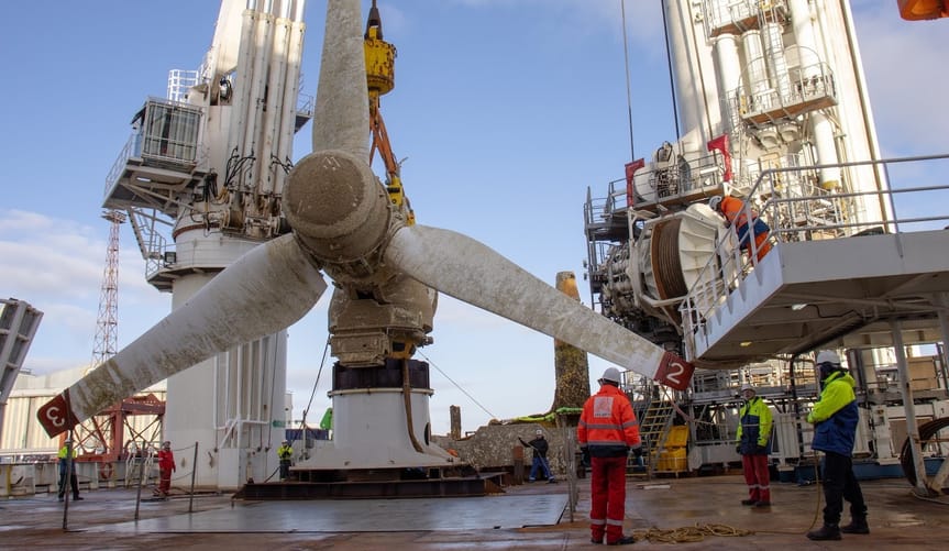 A white three-bladed turbine sits on a dock as workers in bright safety jackets stand nearby