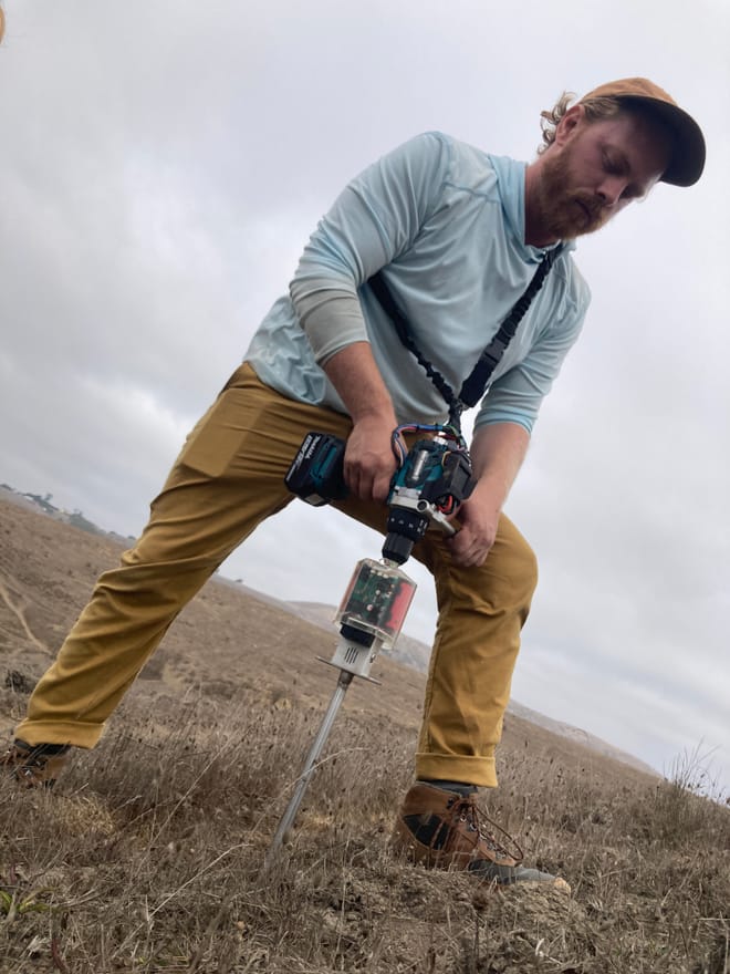 A man with red hair, a blue shirt, and khaki pants plunges the tip of a long metal soil probe into a field to take a sample