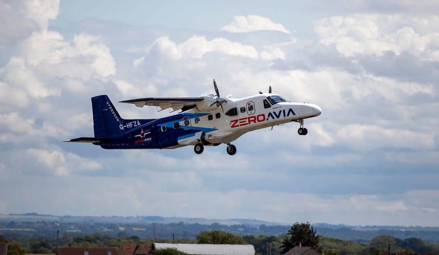 A blue and white propeller aircraft flies over a green countryside