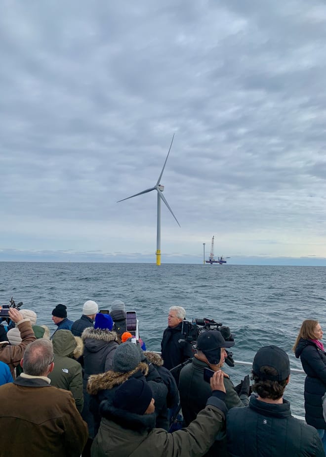 A group of people stand on the deck of ship with cellphones. A wind turbine is seen in the distance.