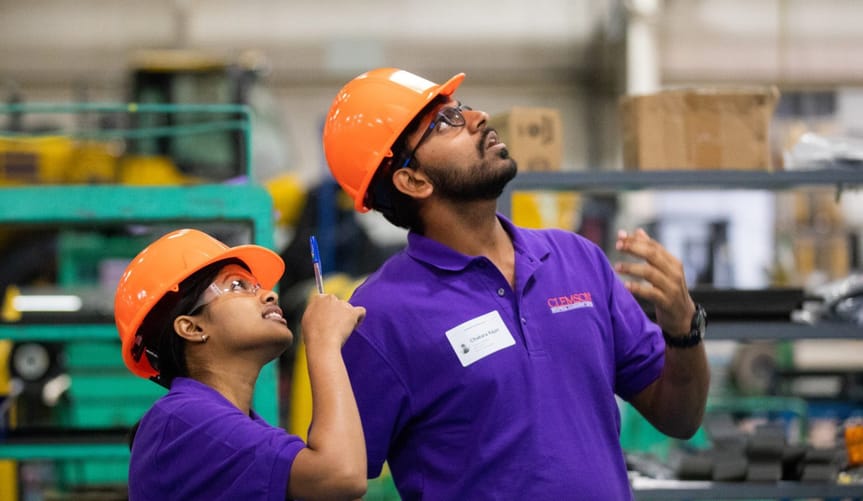 Two students of color with black hair and orange hard hats look up during a free energy assessment of a factory.