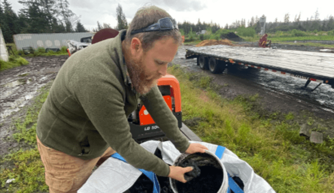A man with light skin and strawberry blonde hair and beard holds up a bucket of black organic matter on a farm