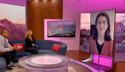 Two news anchors sitting on a blue couch speak to a woman on a video screen with shoulder-length brown hair and pale glasses