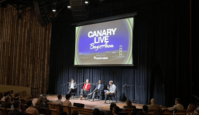 Four people sit in a semicircle on an theater stage under a large projection screen that says Canary Live Bay Area
