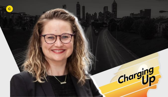 A smiling woman with light skin and brown-blond hair wearing glasses and a black blouse. A graphic overlay says CHARGING UP