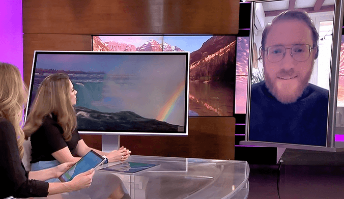 Two women television hosts speak with a man with reddish hair wearing glasses and a black turtleneck via video chat