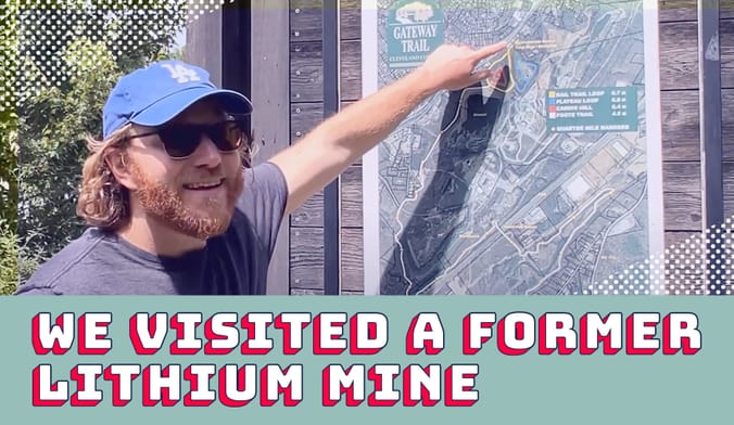 A man with reddish hair wearing a blue baseball cap and sunglasses points at a spot on a trail map display