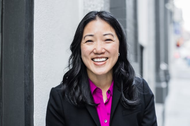 A smiling woman with black hair wearing a pink shirt and black jacket