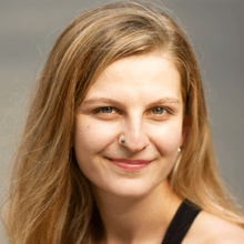 A smiling woman with a light skin tone, long blonde hair and a nose stud wearing a black sleeveless shirt