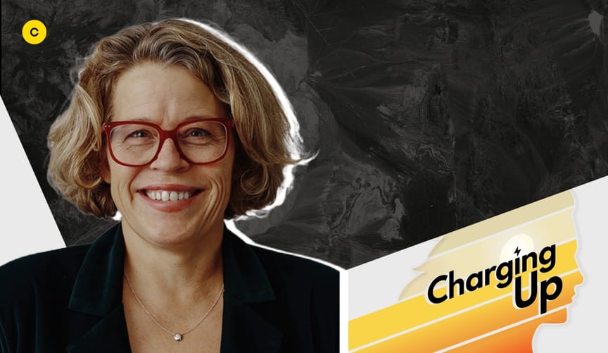 A white woman with short blonde hair and red glasses wearing a black blazer next to a graphic that reads charging up