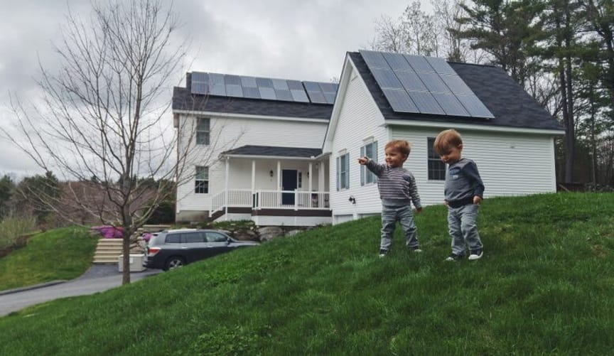Two small toddler boys with red hair stand on a lawn in front of a white house with solar panels on the roof